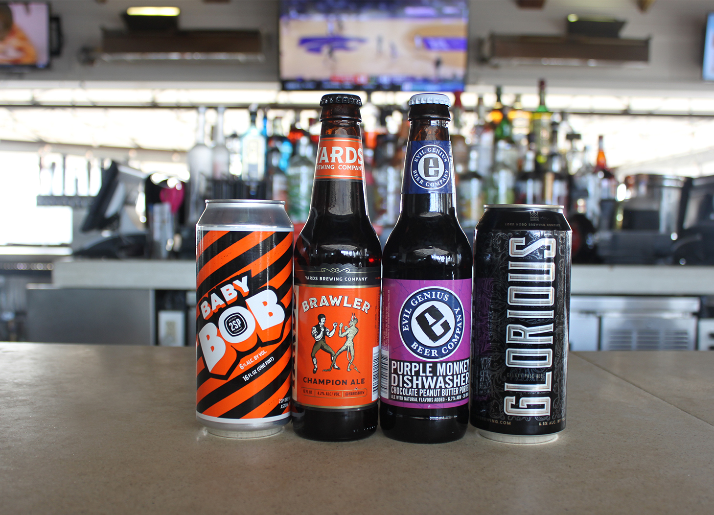 Four craft beers with Tavola bar in the background including an orange and black can of Baby Bob, an orange labeled bottled of Yards Brawler, a purple labeled bottle of Even Genius Purple Monkey and a black and sliver can of Glorious beer.