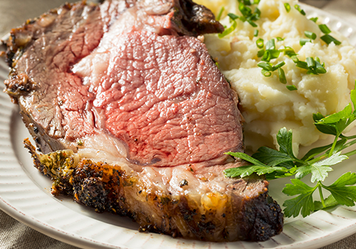 Queen cut prime rib of beef cooked and served on a white dinner plate next to masked potatoes garnished with green herbs.