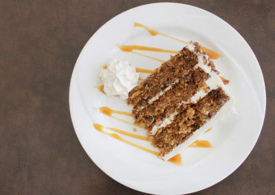 Slice of three-layer carrot cake with white icing on a white plate garnished with caramel drizzle and whip cream.