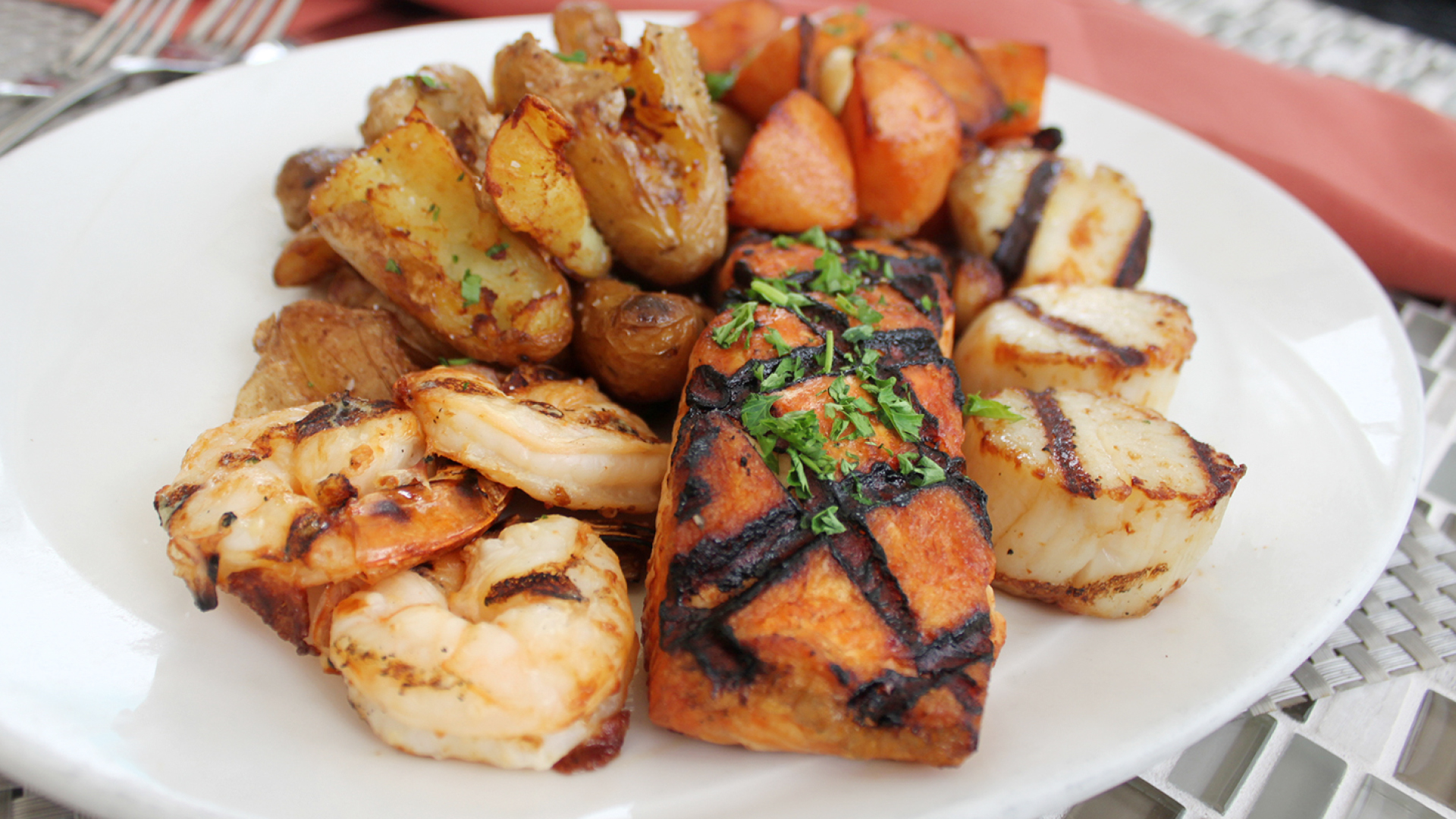 Seafood dinner on a white plate with grilled shrimp, grilled salmon filet and grilled scallops with roasted fingerling potatoes and roasted carrots.