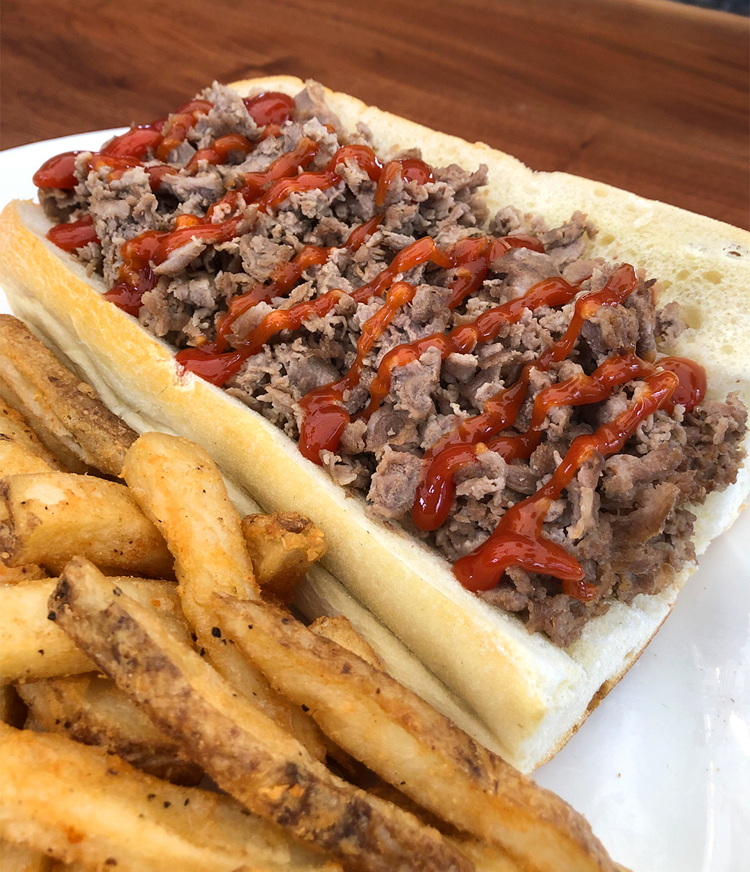 Cheesesteak with ground meet and drizzled with ketchup on a long roll on a white plate next to french fries.