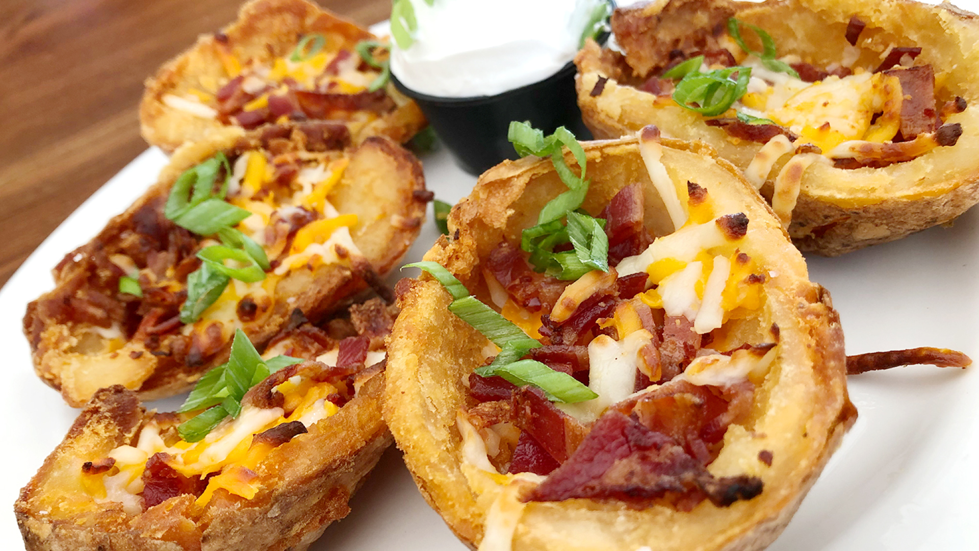 Five potato skins topped with melted shredded cheese, bacon pieces and green scallions on a white plate with a small soufflé cup of sour cream.