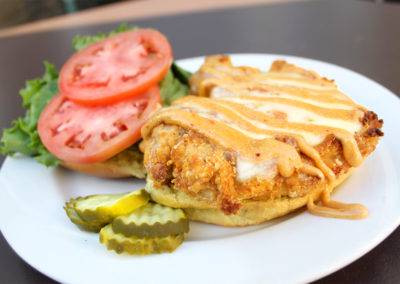 Open faced sandwich with piece of fried chicken, melted cheese and chipolte sauce drizzle alongside sliced tomato and lettuce with side of pickles on a white plate.