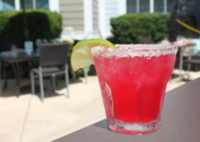 Bright pink colored margarita in a rocks glass with salted rim and lime wedge garnish with outdoor dining tables in background.