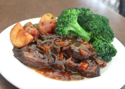 Tender beef short ribs slow cooked in a red wine vegetable sauce on a white circular plate with broccoli florets and roasted potato wedges.