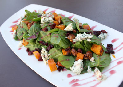 Fresh spinach salad with roasted diced sweet potatoes, Danish blue cheese crumbles, candied pecans, and craisins in a blueberry pomegranate dressing on a white rectangular plate.